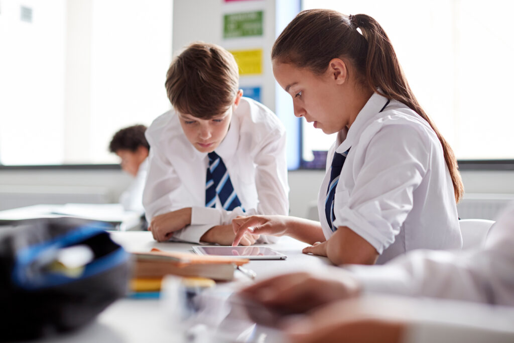 Two High School Students Wearing Uniform Working Together At Desk Using Digital Tablet