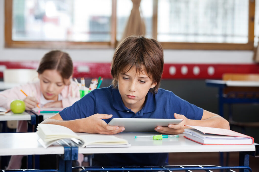 Elementary schoolboy using digital tablet with classmate studying in background at classroom